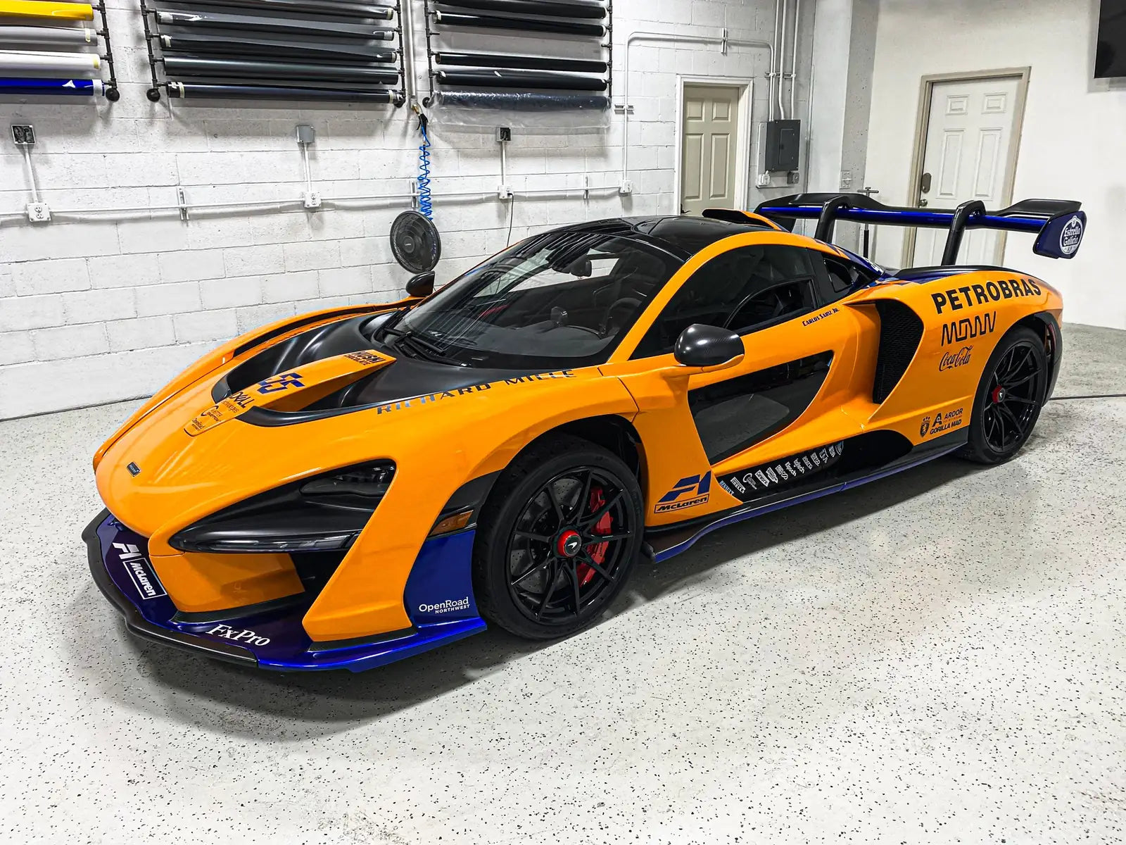 McLaren Senna with a full orange and blue racing livery wrap and sponsor decals, parked inside the Ardor Printing shop