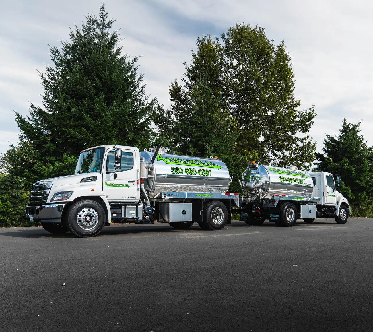 Two white septic tanker trucks with polished stainless tanks, green lettering and phone number graphics parked on asphalt